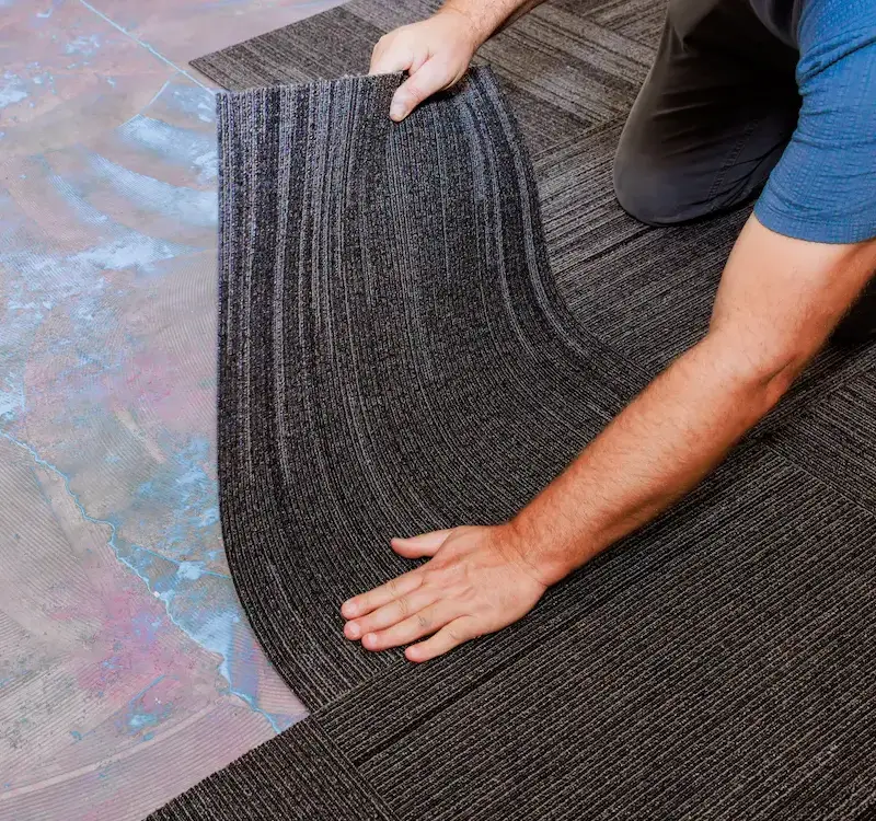 Professional installer placing modular carpet tiles over a pressure-sensitive adhesive on a commercial concrete floor.